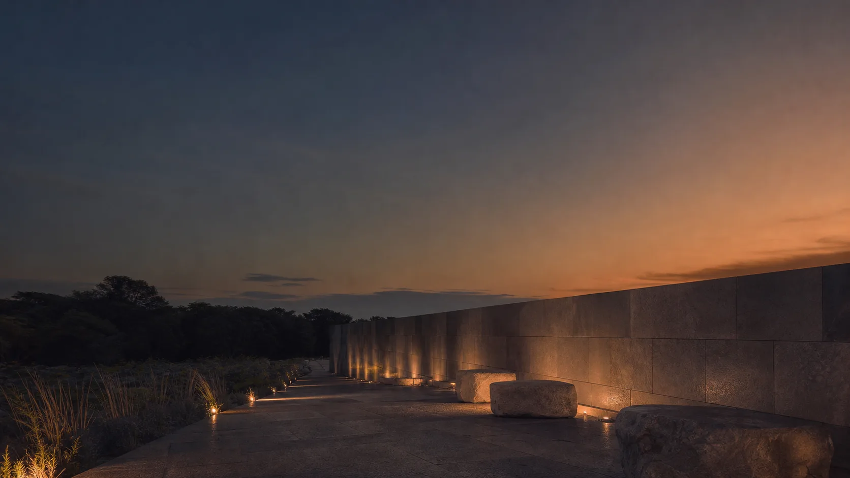 Mur minéral et chemin éclairé dans un lieu de mémoire au crépuscule.